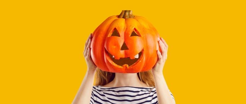 Woman in stripy top against yellow background hiding face behind Halloween pumpkin lantern.