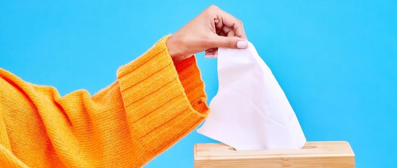 Woman in orange jumper with tissue box against blue background.