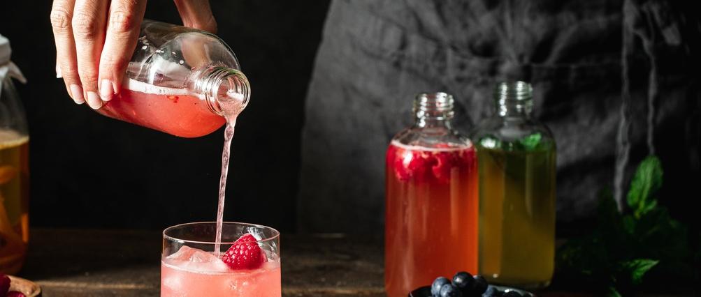 Hand pouring pink raspberry kombucha from bottle into glass with ice, with colourful drink bottles in background.