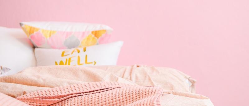 Pink, white and yellow bed covers, with 'Eat Well' printed on pillow. Set against pink background.