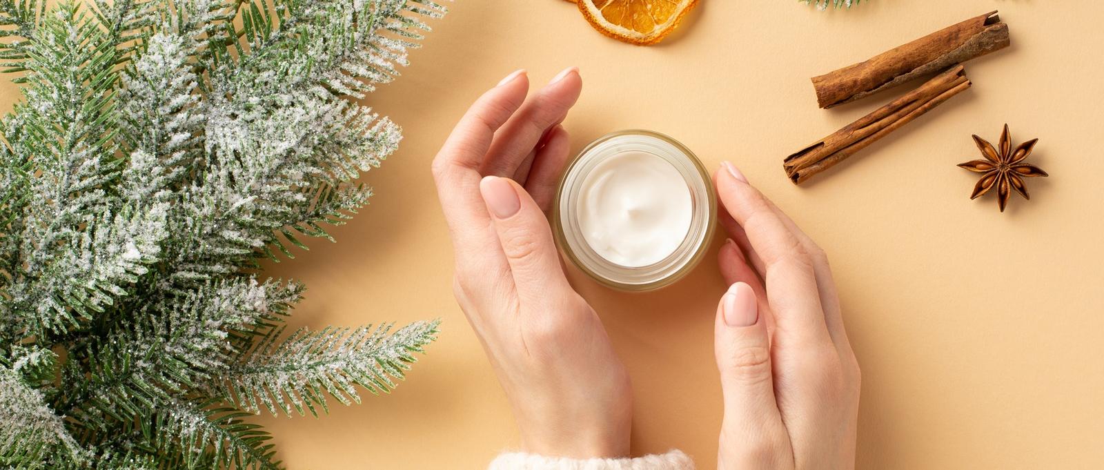 Winter skin care cosmetics concept. First person top view photo of girl's hands in pullover cream jar spruce branches dried orange slices cinnamon sticks and anise on isolated pastel beige background