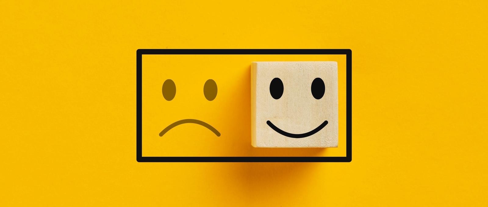 Two wooden cubes with happy mood icon over sad mood icon on a yellow background
