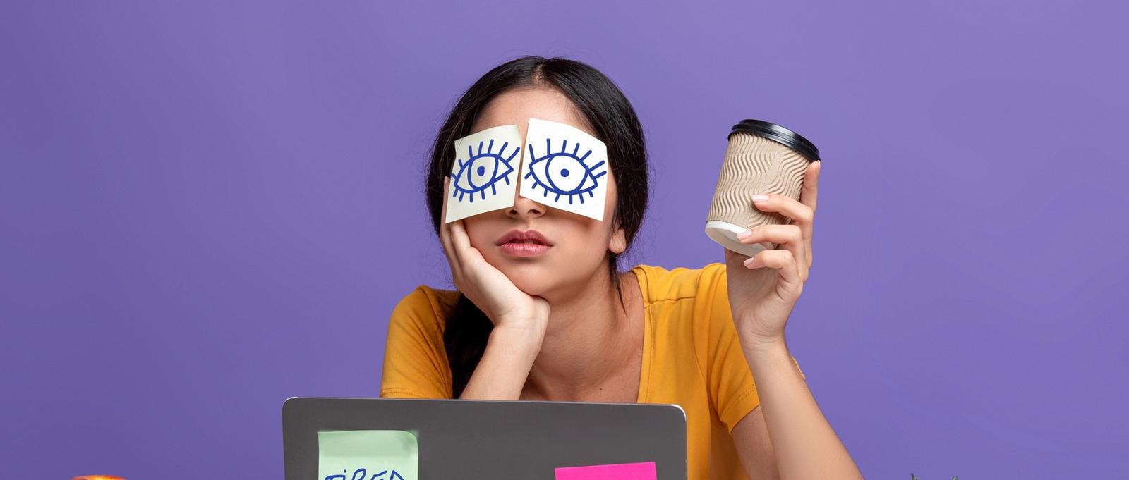 Portrait of exhausted young woman sitting at workplace with stickers on eyes and holding cup with coffee, working on laptop at table over purple studio background