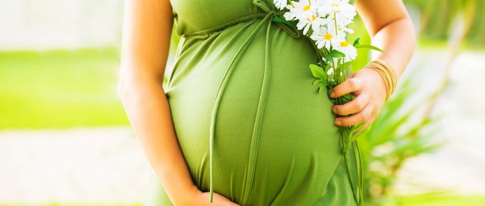 Pregnant woman in green dress holding white daisies against her belly in a sunny outdoor setting.