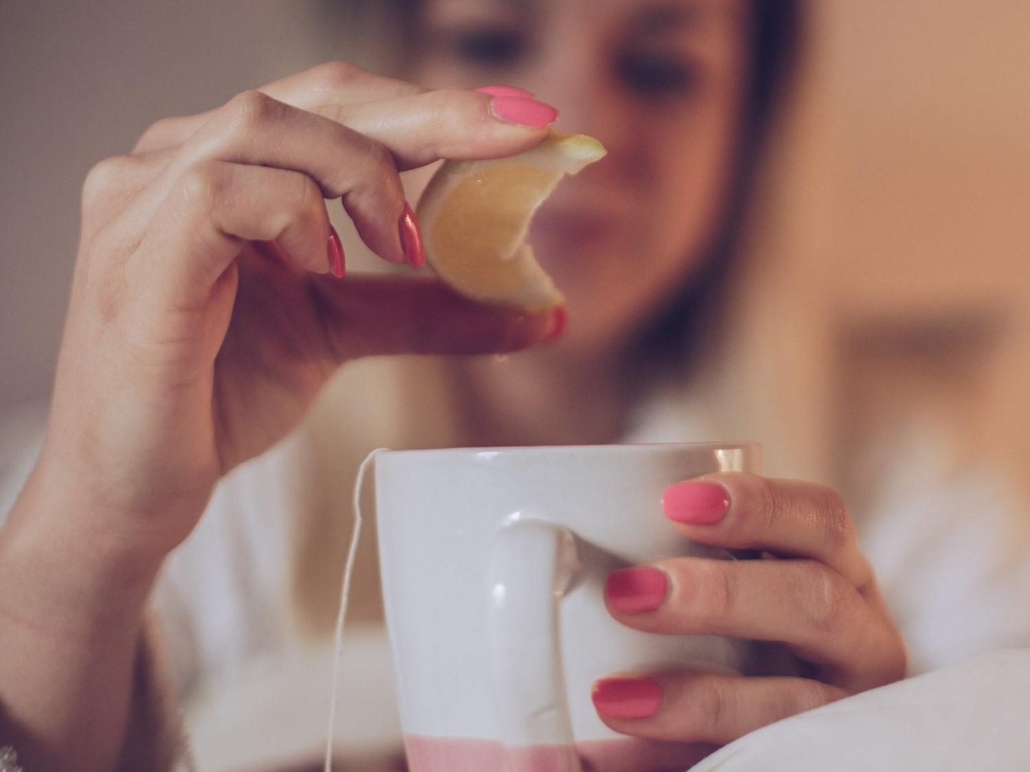 Person with pink manicured nails squeezing lemon into a white teacup filled with tea, background blurred.