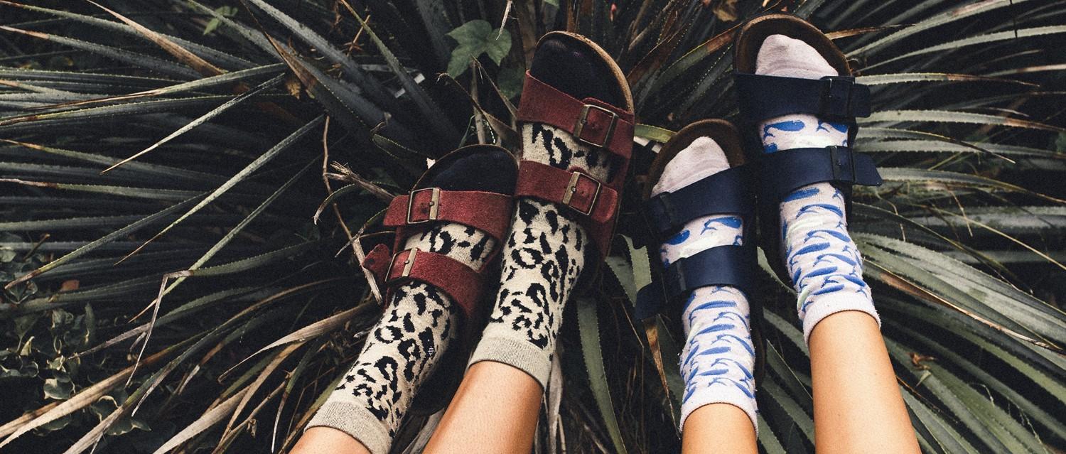 Two pairs of feet wearing sandals with patterned socks - leopard print with brown straps and dolphin print with blue straps.
