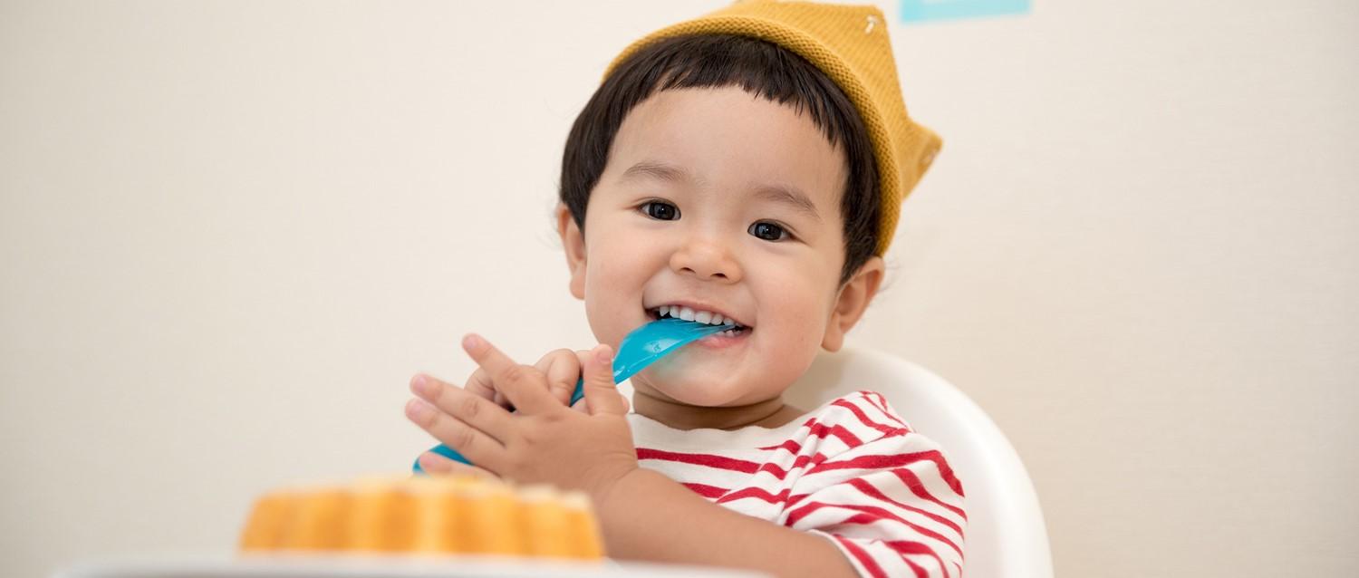 Smiling toddler wearing yellow crown and striped shirt, eating with blue spoon in high chair with food visible.