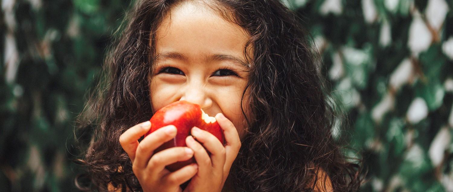 Young child with curly hair smiling while taking a bite from a red apple, green foliage background.