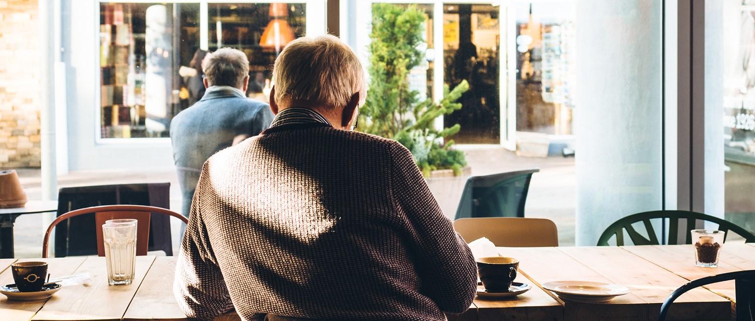 Two elderly people sitting in a bright café with coffee cups on wooden tables, looking out through large windows.