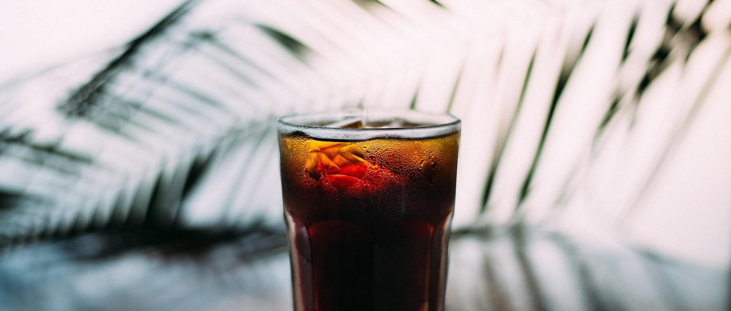 Cold glass of cola with ice cubes and condensation against a blurred background with palm leaf shadows.