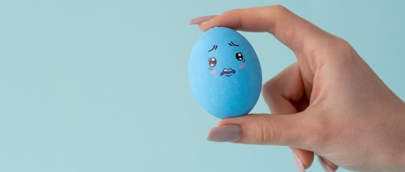 Hand holding sad-faced blue hard boiled egg against blue background.