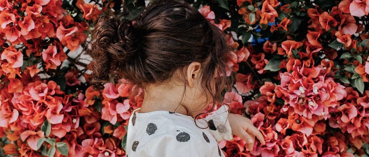 Person with dark hair in a bun wearing polka dot clothing surrounded by vibrant coral flowers and green leaves.