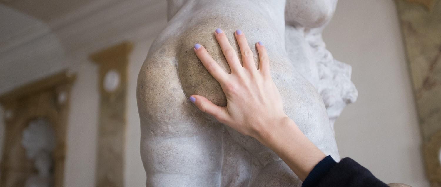 Hand with lavender nail polish touching a classical marble statue in a museum gallery.