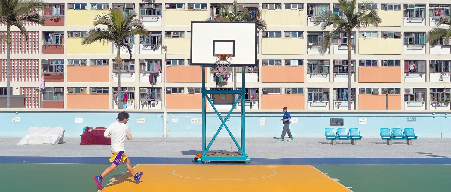 Basketball court with colourful apartment building backdrop, palm trees, and people walking on the court.