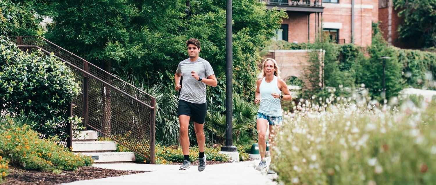 Two runners jogging along a garden path with lush greenery and brick buildings in the background.