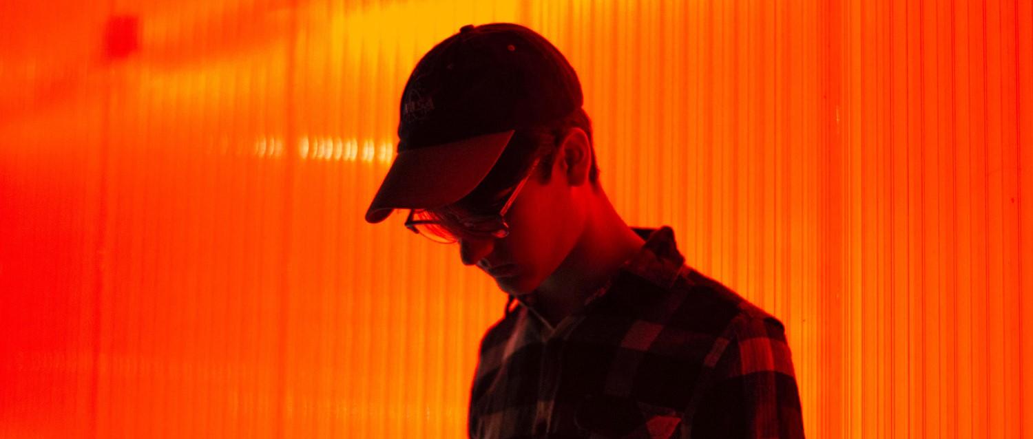 Silhouette of person wearing cap and glasses against vibrant orange background with vertical light streaks.