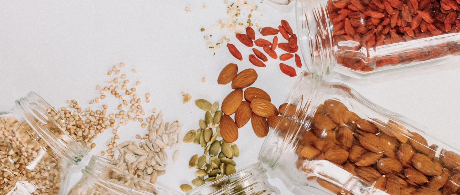 Glass jars spilling various nuts, seeds and goji berries on white background, showcasing healthy snack ingredients.