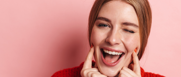 Person with auburn hair wearing red sweater laughing joyfully against pink background